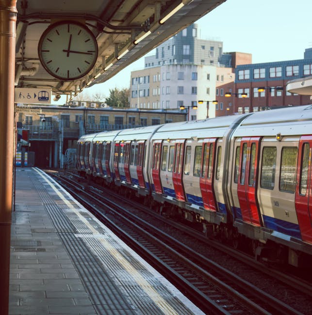 View of a train station platform adjacent to railway tracks, with a brick wall and white metal fencing separating the platform from a residential area. Tall modern apartment buildings are visible in t