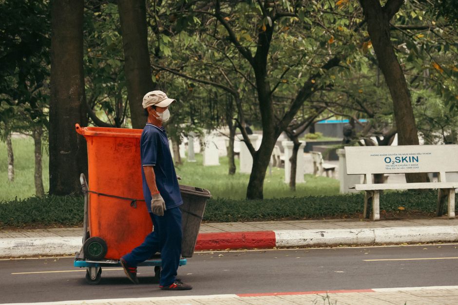 A street cleaner wearing a beige cap, white face mask, navy blue uniform with orange accents, and grey gloves is pushing a trolley with an orange waste bin and a smaller brown bin along a sidewalk. Th